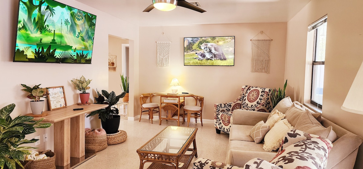 A comfortable living space features a light color palette with decorative plants and woven accents. A large television hangs on the wall, opposite a cozy sofa with patterned cushions. A dining table with chairs is positioned beside the sofa, creating an inviting atmosphere.