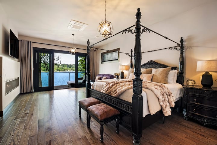 Wake up to expansive lake views in the master bedroom with fireplace, TV, sitting area, private balcony with outdoor daybed, night stands, dresser, and adjacent master bathroom.