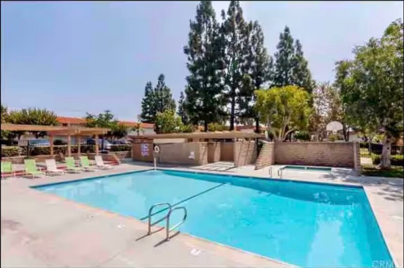 A swimming pool is showcased, surrounded by tall trees and well-maintained green spaces. Lounge chairs are positioned around the pool, providing a place to relax. The shallow end features a railing for easy access, with sunny skies above enhancing the outdoor ambiance.