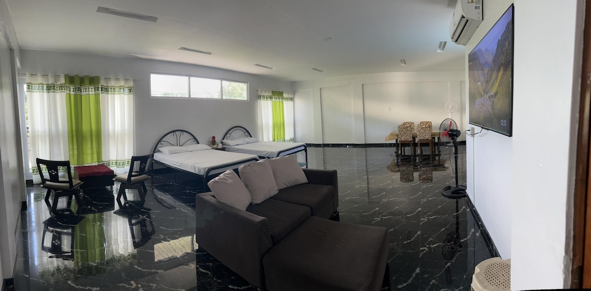 A spacious room features two beds with light-colored linens. A comfortable couch is positioned near a large television on the wall. Natural light enters through sheer green curtains framing the windows. A dining area with two chairs and a small table is situated in the corner.