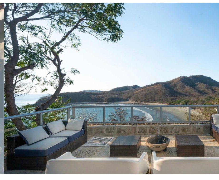 An outdoor seating area offers panoramic views of the coastline and surrounding hills. Comfortable lounge furniture is arranged around a central fire pit, with trees providing partial shade. The clear sky enhances the tranquil ambiance of the setting.