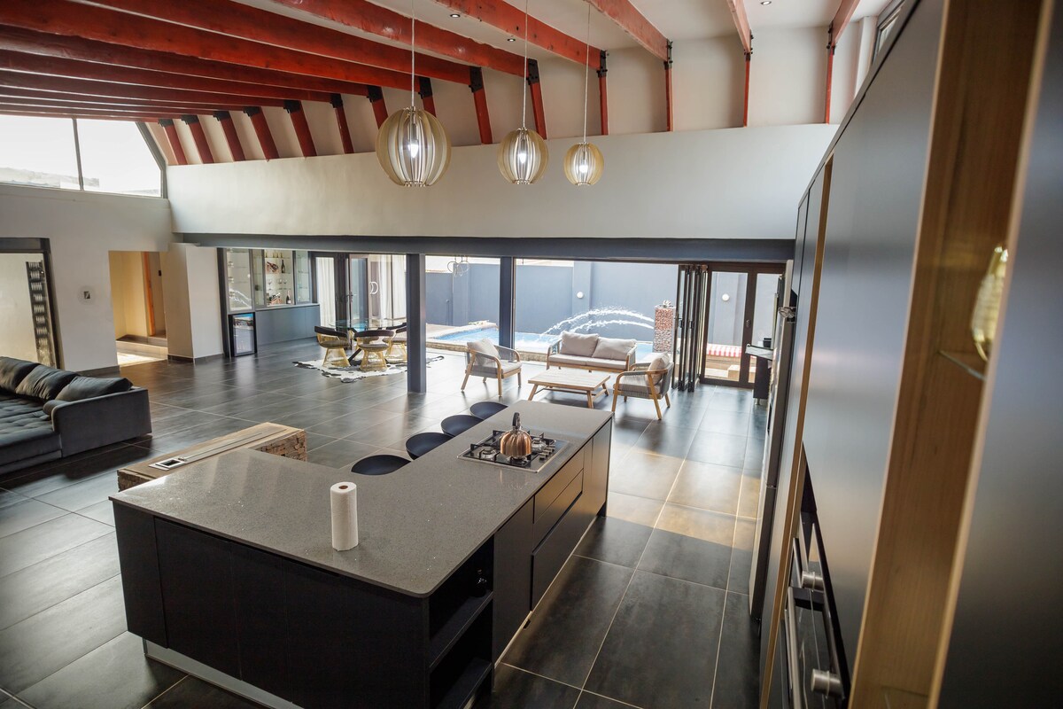 The open-plan kitchen features a spacious island with seating for four, complemented by modern pendant lighting. Floor-to-ceiling windows provide natural light and views of the outdoor entertainment area, blending the interior with the pool area beyond.