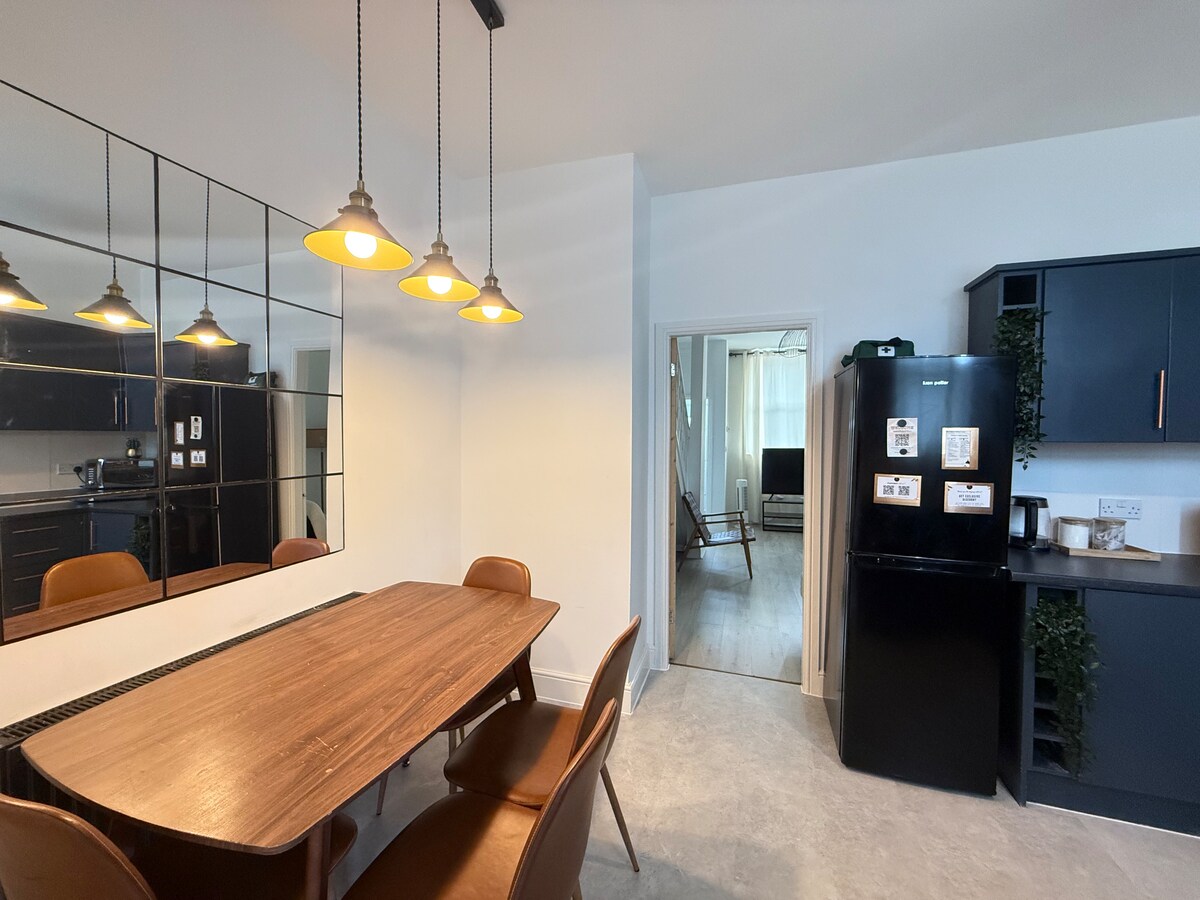 A modern dining area features a wooden table surrounded by stylish chairs, with warm pendant lights overhead. A sleek black refrigerator is positioned beside the kitchen area, reflected in a large mirror that enhances the space's openness.