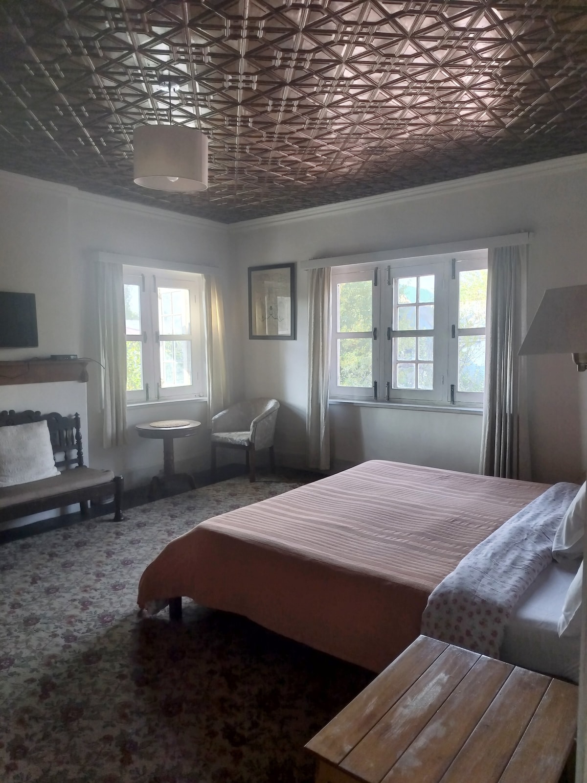 A bright bedroom features a spacious bed covered in soft linens, complemented by a rustic wooden nightstand. A cozy seating area includes a light-colored chair and a small round table. Natural light enters through multiple windows, while textured ceiling tiles add character.
