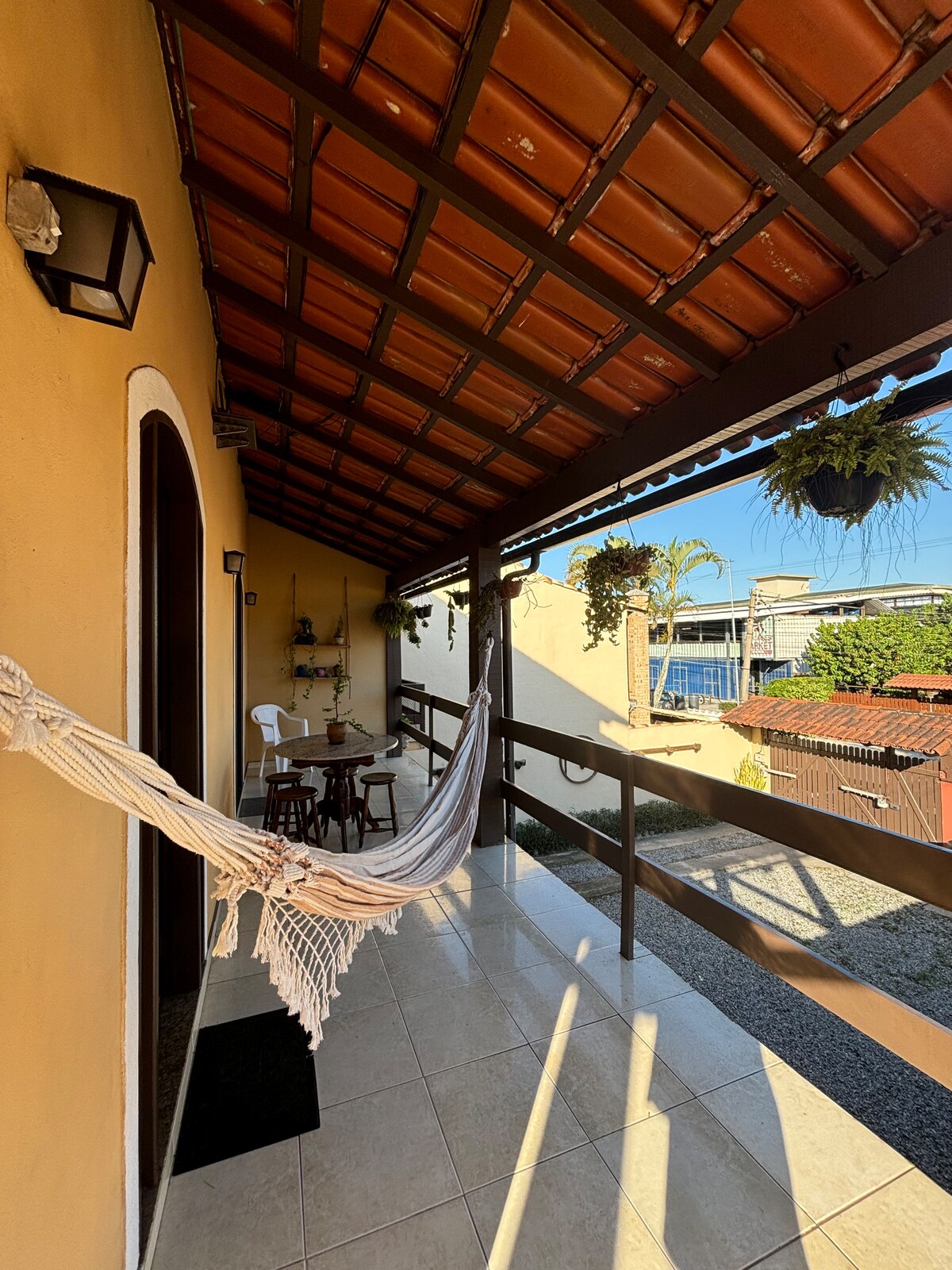 A covered balcony features a hammock and a small wooden table with two chairs. Hanging plants add a touch of greenery. The tiled floor reflects warm tones, and a railing provides safety. A glimpse of the street and surrounding areas can be seen in the background.