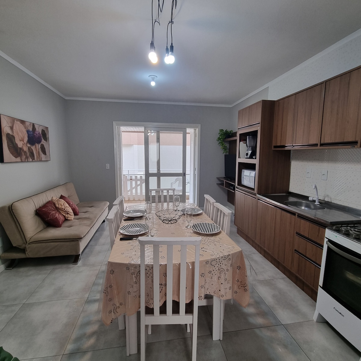 The kitchen and dining area features modern wooden cabinetry and a large dining table set for six. A light-colored sofa is positioned nearby, enhancing the open layout. Natural light enters through sliding glass doors that lead to a small balcony.