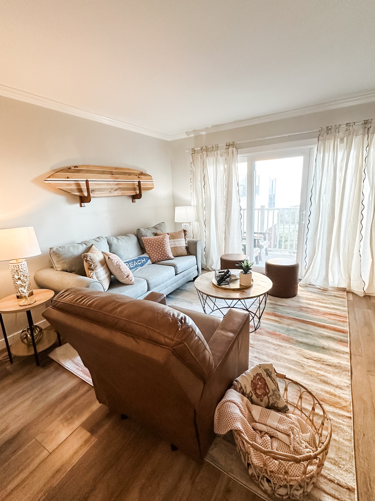 A cozy living area is equipped with a light gray sofa and an armchair, accompanied by a wooden coffee table. Natural light enters through large sliding doors, revealing outdoor views. A decorative surfboard is mounted on the wall, while textured rugs add warmth to the space.
