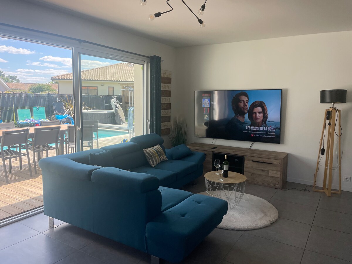 A comfortable living area is highlighted by a blue sectional sofa facing a large flat-screen television. Natural light fills the space through sliding glass doors, which open to a deck with outdoor seating and a pool view. A round coffee table adds to the room's functionality.