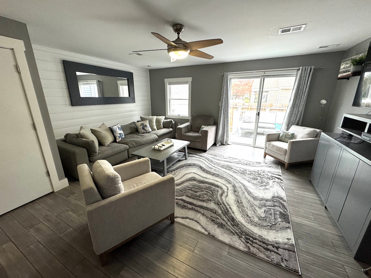 The living area features comfortable seating arrangements, including a sofa and two armchairs, situated on a stylish area rug. A ceiling fan adds airflow, while large sliding doors provide access to the outdoor deck, allowing natural light to brighten the space.