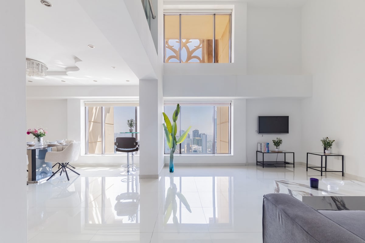 An open and airy living space is presented, featuring large windows that provide panoramic views of the surrounding area. A sleek gray sofa is positioned in front of a modern television, while a dining area with stylish chairs is set beside a tall indoor plant.