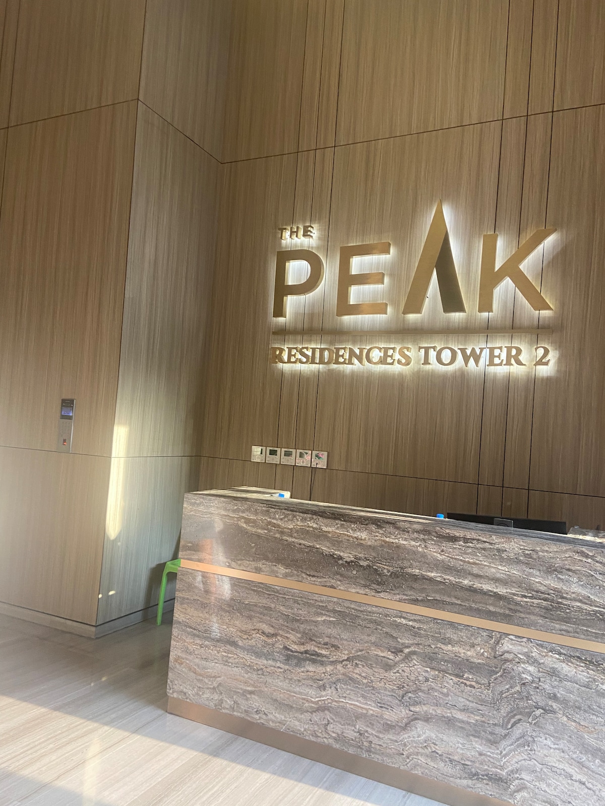 The reception area features a modern design with warm wooden paneling and a sleek stone front desk. A lit sign displaying 'THE PEAK RESIDENCES TOWER 2' is prominently displayed, indicating the location. Natural light filters in, enhancing the warm tones of the space.
