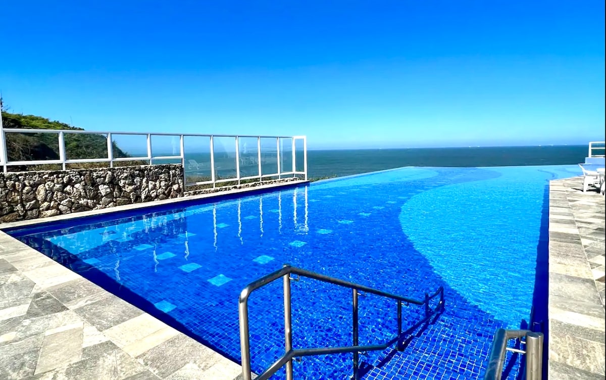 An expansive infinity pool is shown against a clear blue sky, offering stunning views of the ocean. The pool area features a stone wall and a set of stairs leading into the water, highlighting the serene setting.