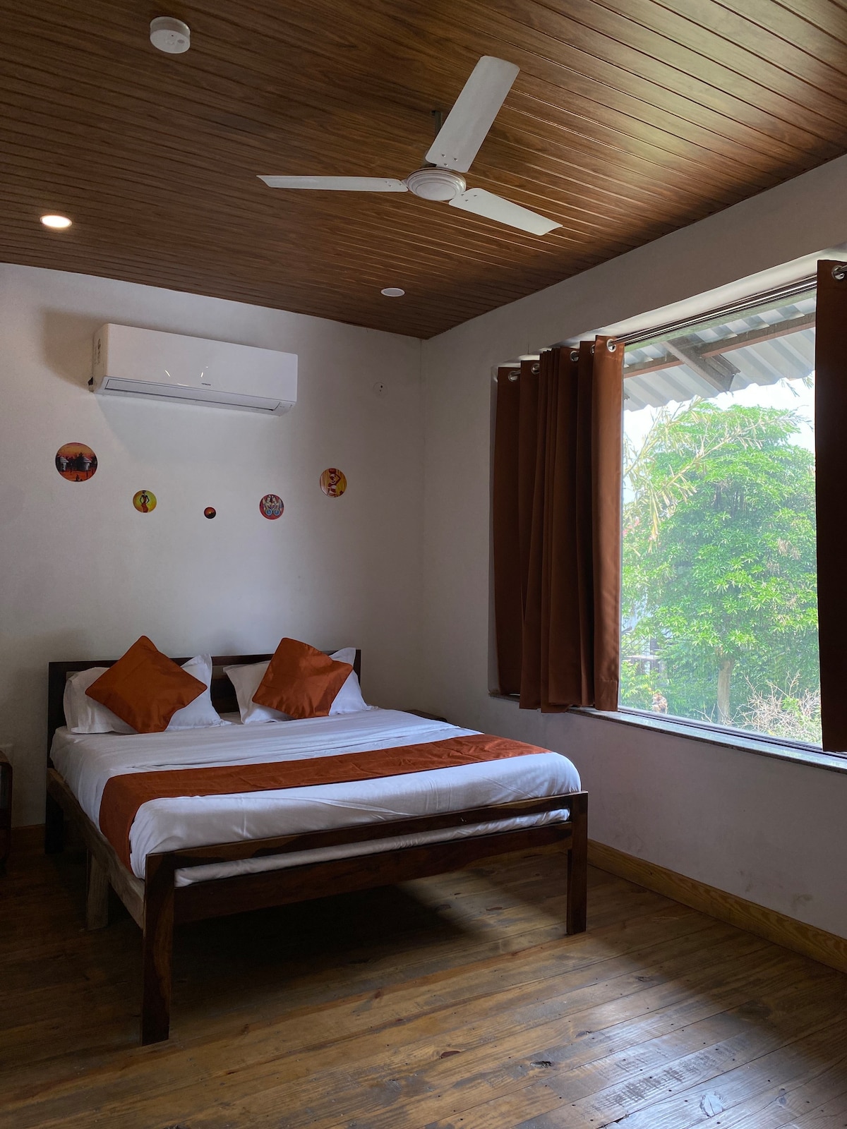 A bedroom features a wooden bed with white linens and orange accent pillows. Natural light enters through a large window, framed by brown curtains. A ceiling fan is mounted above, and decorative wall art adds a touch of character.