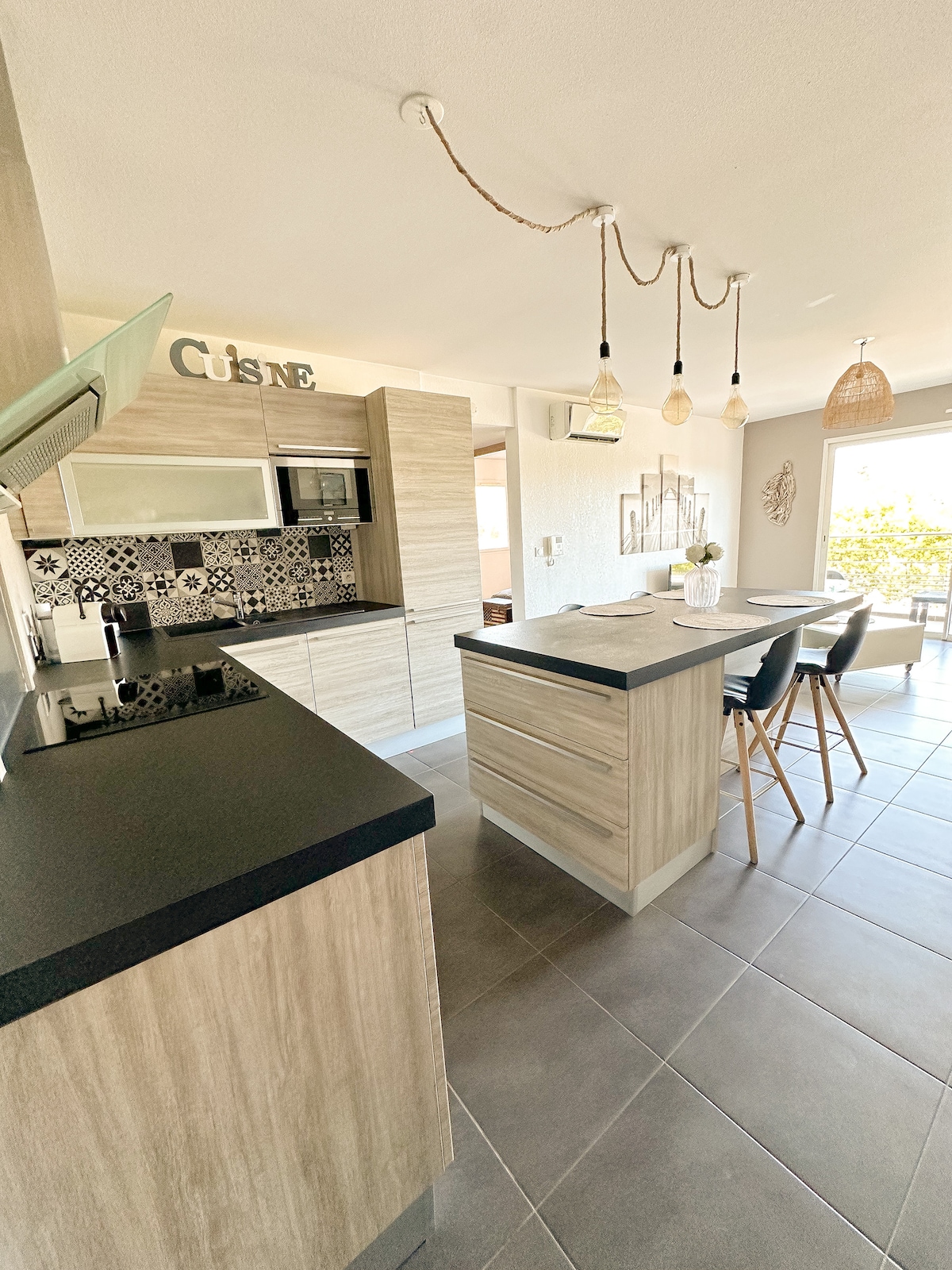 The kitchen features contemporary cabinetry with a natural wood finish and black countertops. A spacious island serves as a dining area with three high stools. Decorative elements include a patterned backsplash and pendant lighting, while large windows allow natural light to illuminate the space.