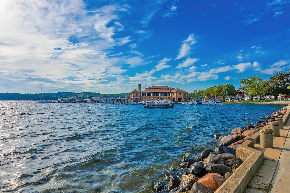 A serene waterfront view depicts a calm lake with gentle ripples under a bright blue sky adorned with scattered clouds. The shore features a mix of rocks and a paved walkway, alongside a charming building that offers access to various water activities.
