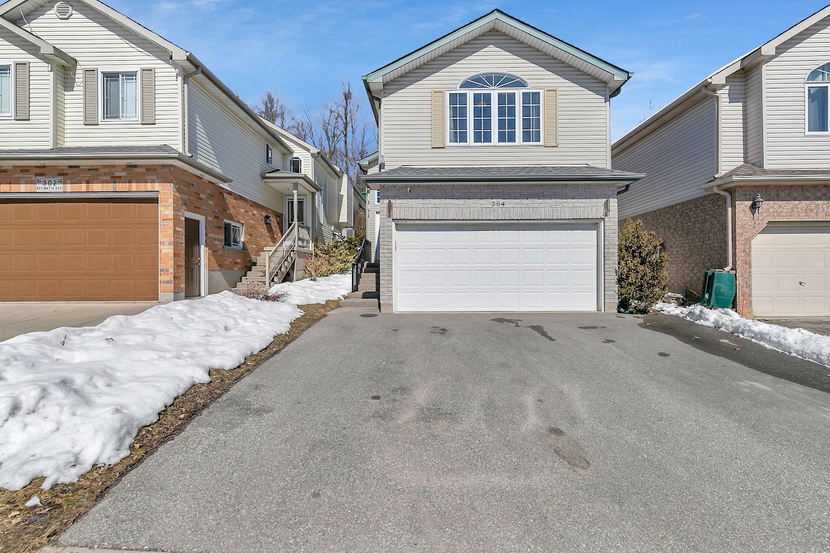 A well-maintained driveway leads to a two-car garage beneath a two-story house. Snow patches are visible on the sides, indicating the transition from winter. The house features an arched window, while neighboring houses display a similar architectural style, creating a cohesive neighborhood appearance.