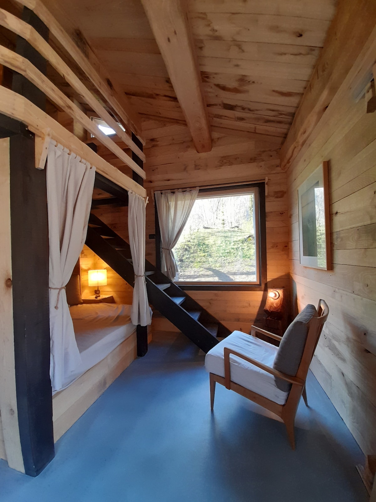 A cozy interior showcases wooden walls and ceiling beams, featuring a sleeping loft accessed by a dark staircase. Natural light filters through a large window, adorned with soft curtains. A stylish chair is positioned nearby, creating a comfortable seating area.