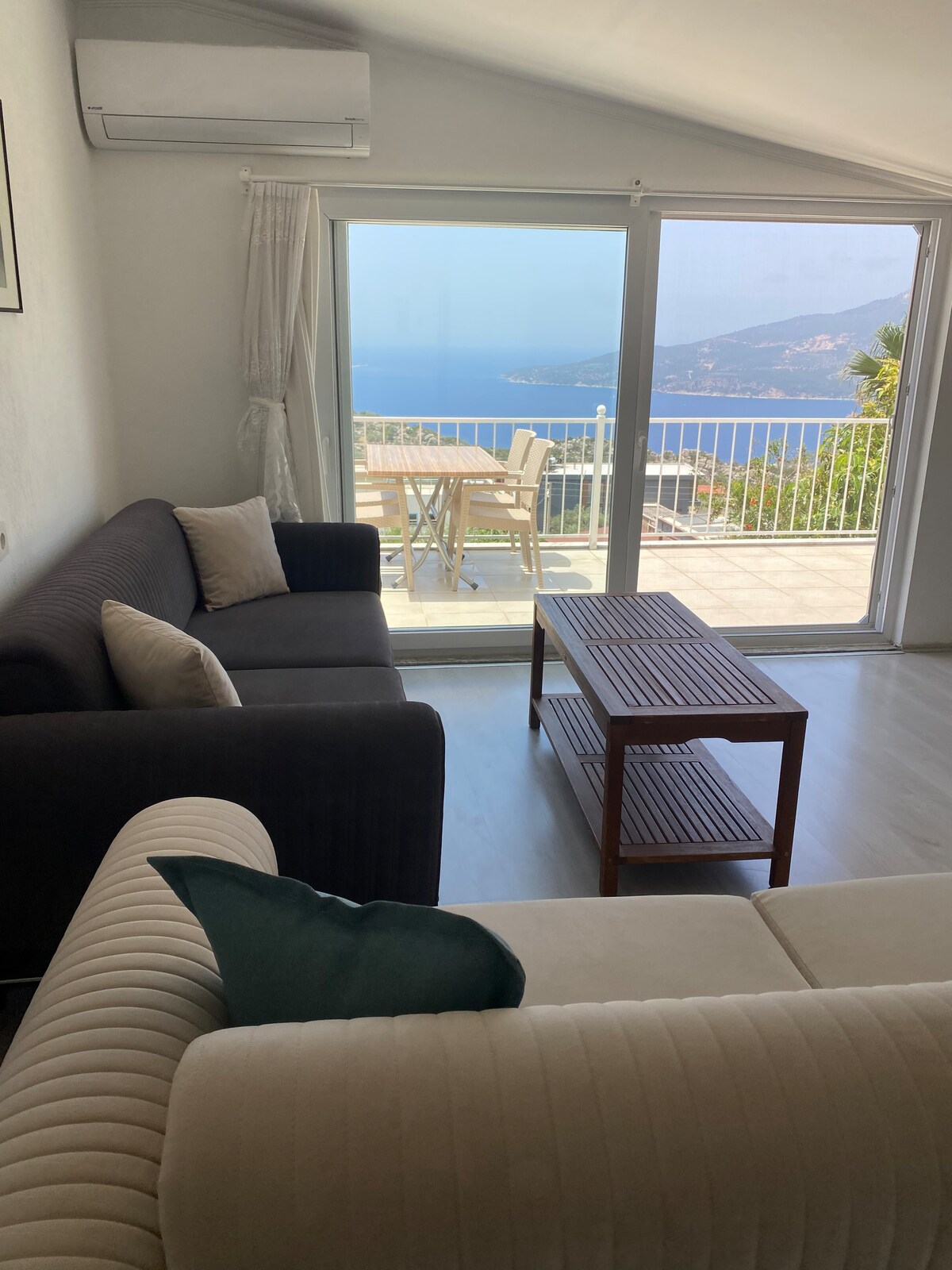 A comfortable sitting area is visible, featuring two sofas and a wooden coffee table. Large glass doors open to a balcony with a table and chairs, offering views of Kalkan Bay and surrounding mountains.