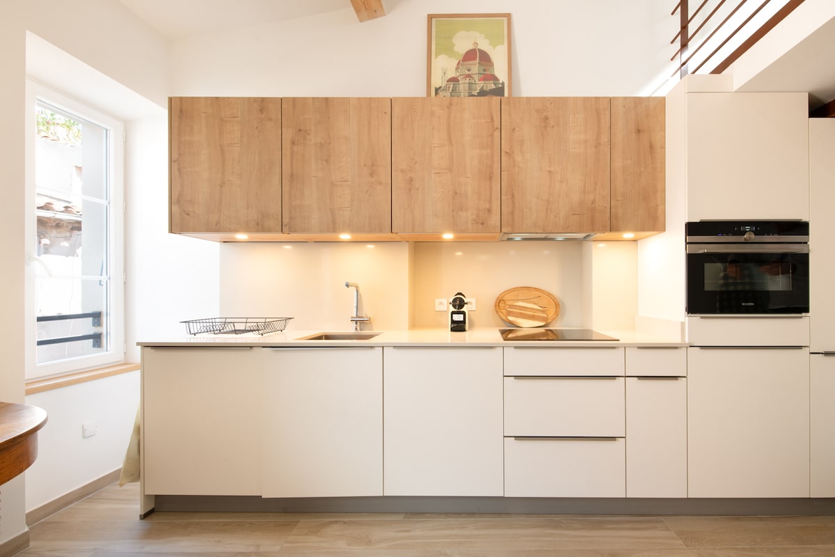 A modern kitchen is presented with light wood cabinetry and a sleek countertop. Under-cabinet lighting illuminates the space, highlighting a few kitchen appliances and utensils. Natural light enters through a window, enhancing the clean and contemporary design.
