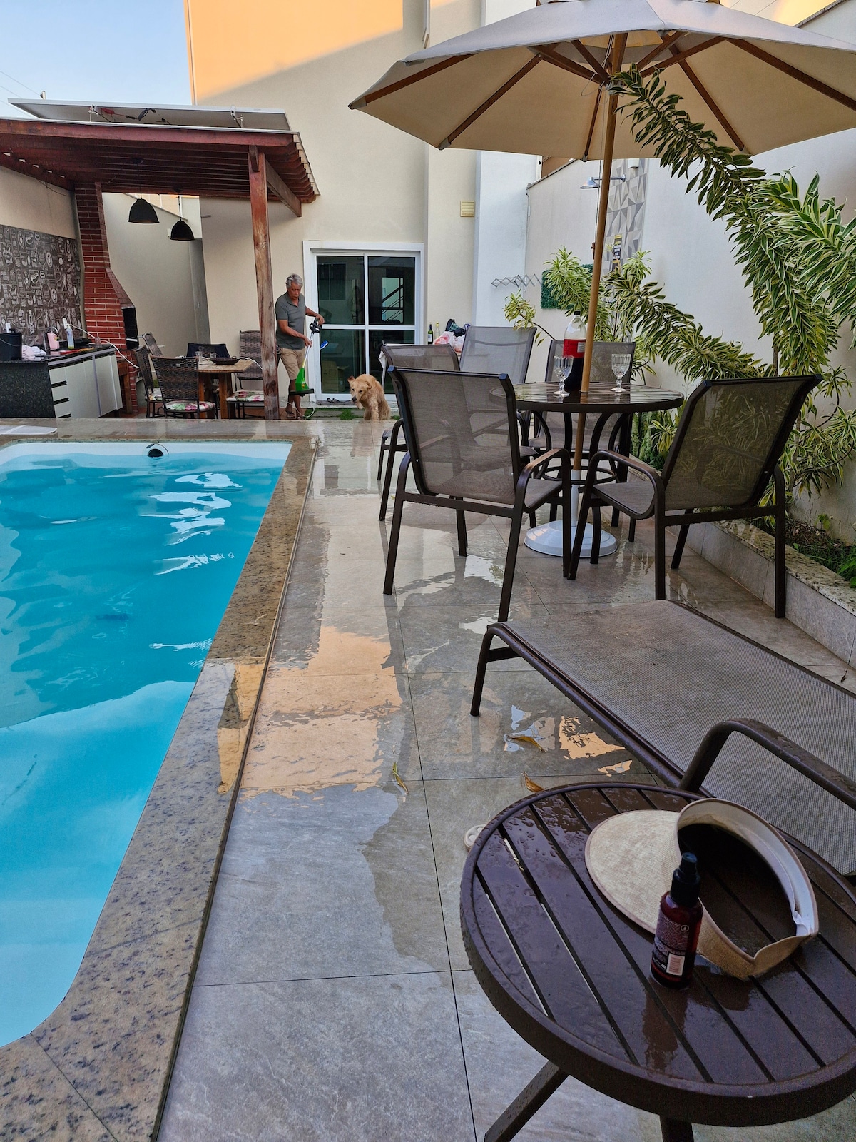 A private pool area is visible, featuring elegant lounge chairs and a table shaded by an umbrella. The pool reflects the sunlight, and a portion of a gourmet outdoor kitchen is seen in the background. Lush greenery surrounds the space, adding a serene touch.