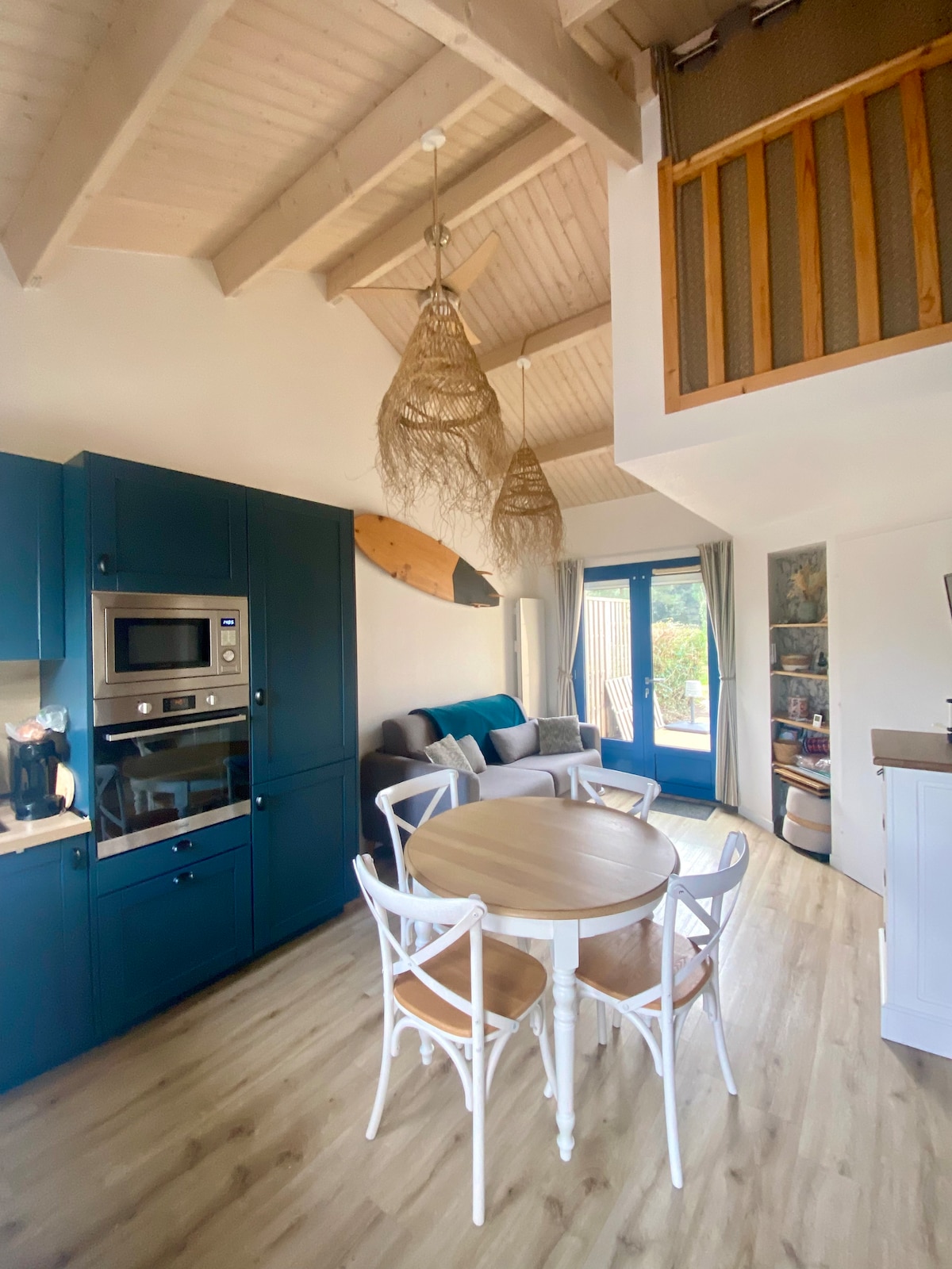 A modern kitchen area features blue cabinetry paired with a circular dining table and four white chairs. A spacious sofa is visible in the corner, accentuated by decorative pillows. Natural light filters through glass doors, revealing outdoor access and enhancing the airy ambiance.