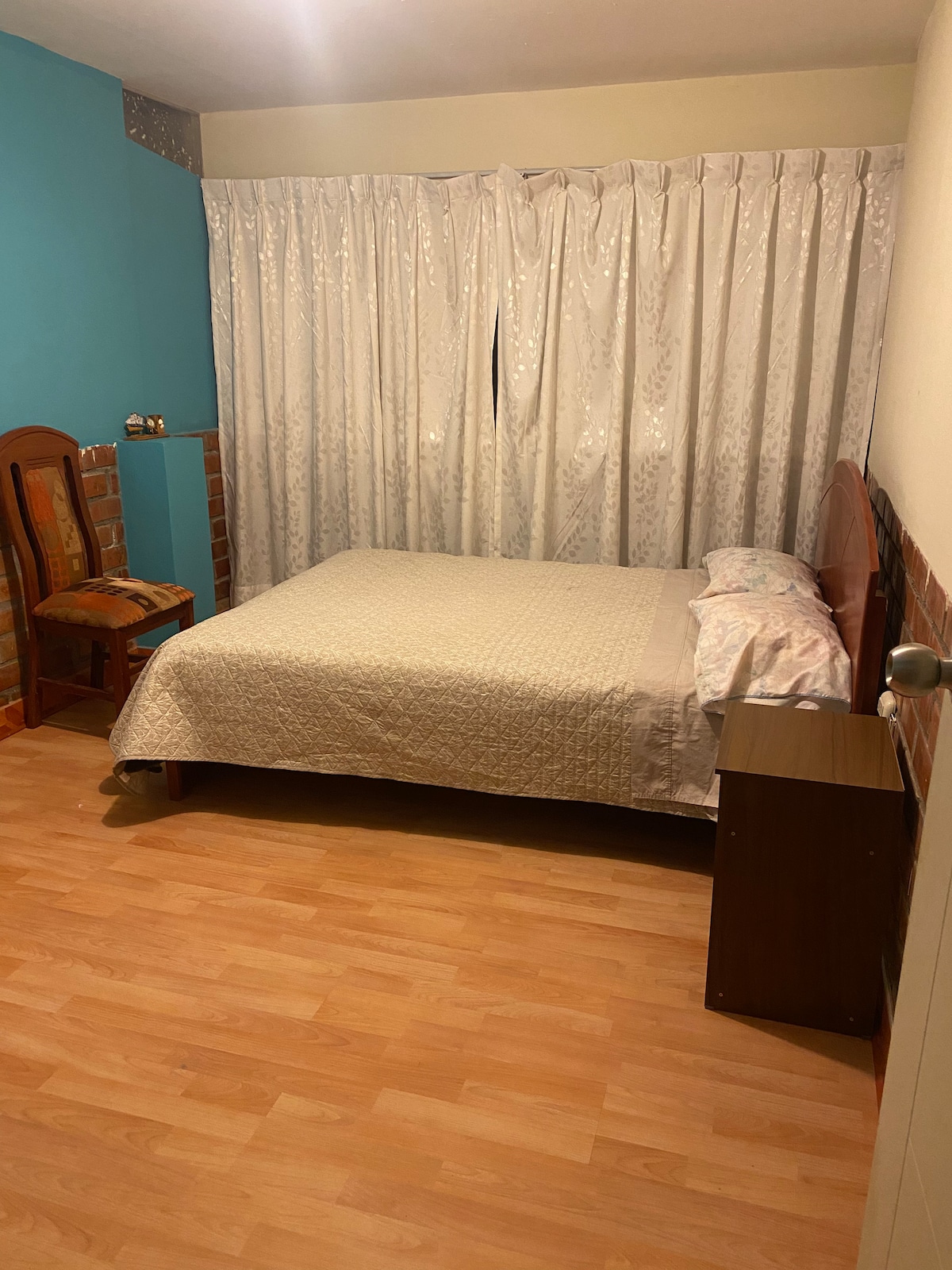 A private bedroom is displayed with a wooden bed covered by a light quilt. A small nightstand is positioned next to the bed. A vibrant blue wall contrasts with the soft beige curtains, allowing natural light to brighten the space. A modest wooden chair sits in the corner.