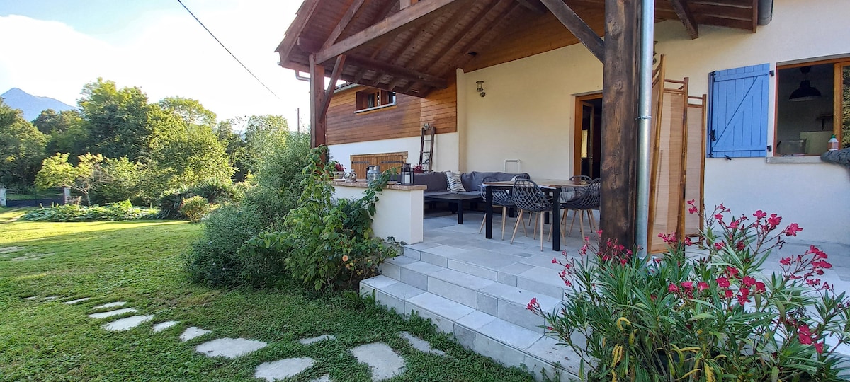 A covered terrace is observed, featuring a wooden ceiling and stone flooring. Outdoor seating is arranged around a table, with potted plants and flowers lining the edges. A well-kept garden is visible in the background, adding to the natural surroundings.