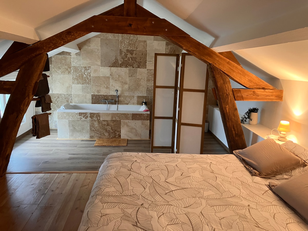 A spacious loft bedroom features exposed wooden beams and light-colored walls. A cozy bed with a patterned duvet is positioned next to a large, stylish bathtub that is set against a feature wall of textured stone tiles. Soft lighting is provided by a bedside lamp.