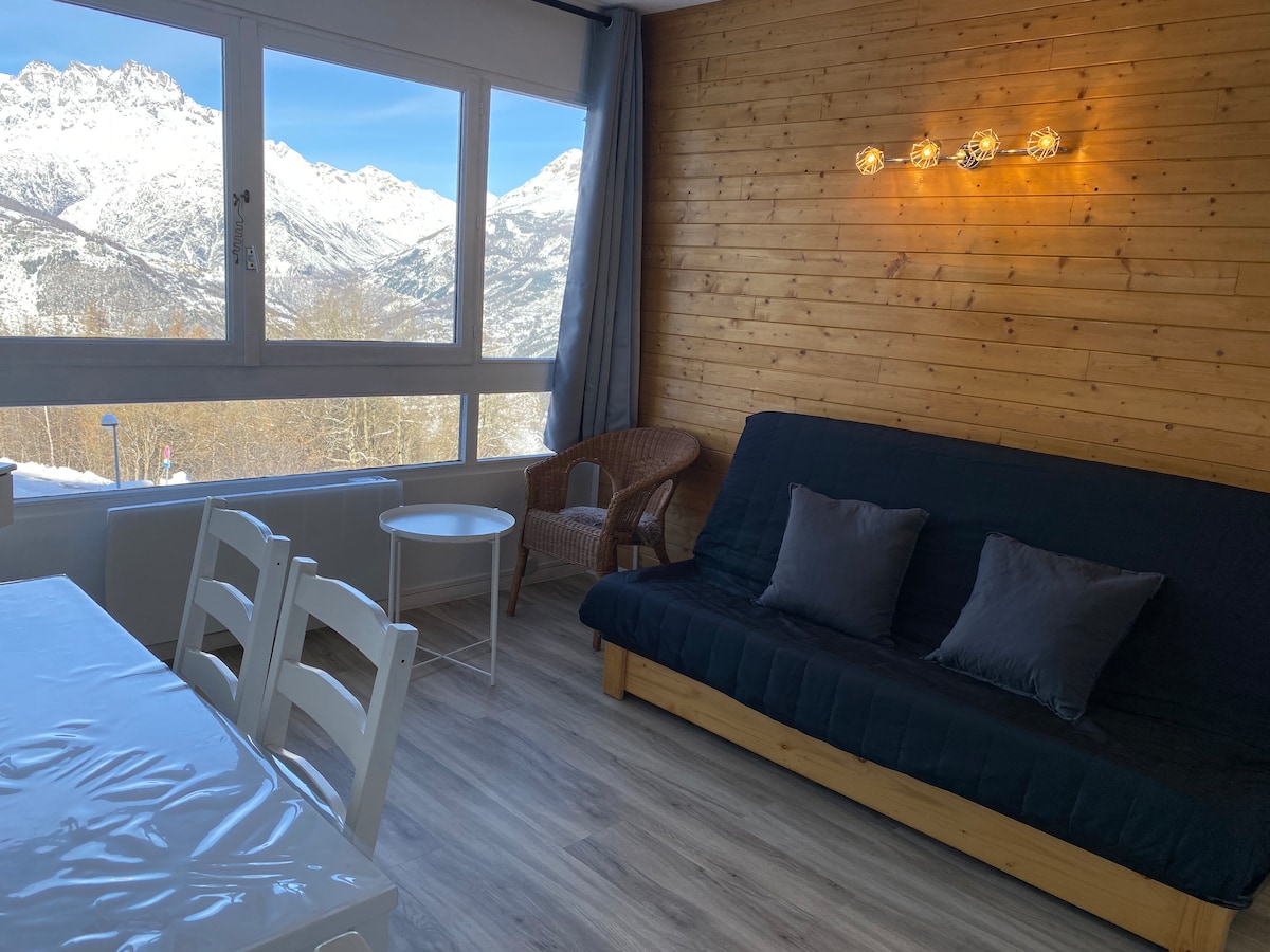 A cozy living area is featured, with large windows offering a scenic view of the surrounding mountains. A comfortable seating area includes a dark sofa bed with pillows and a woven chair. A dining table with white chairs is positioned nearby against a warm wooden wall.