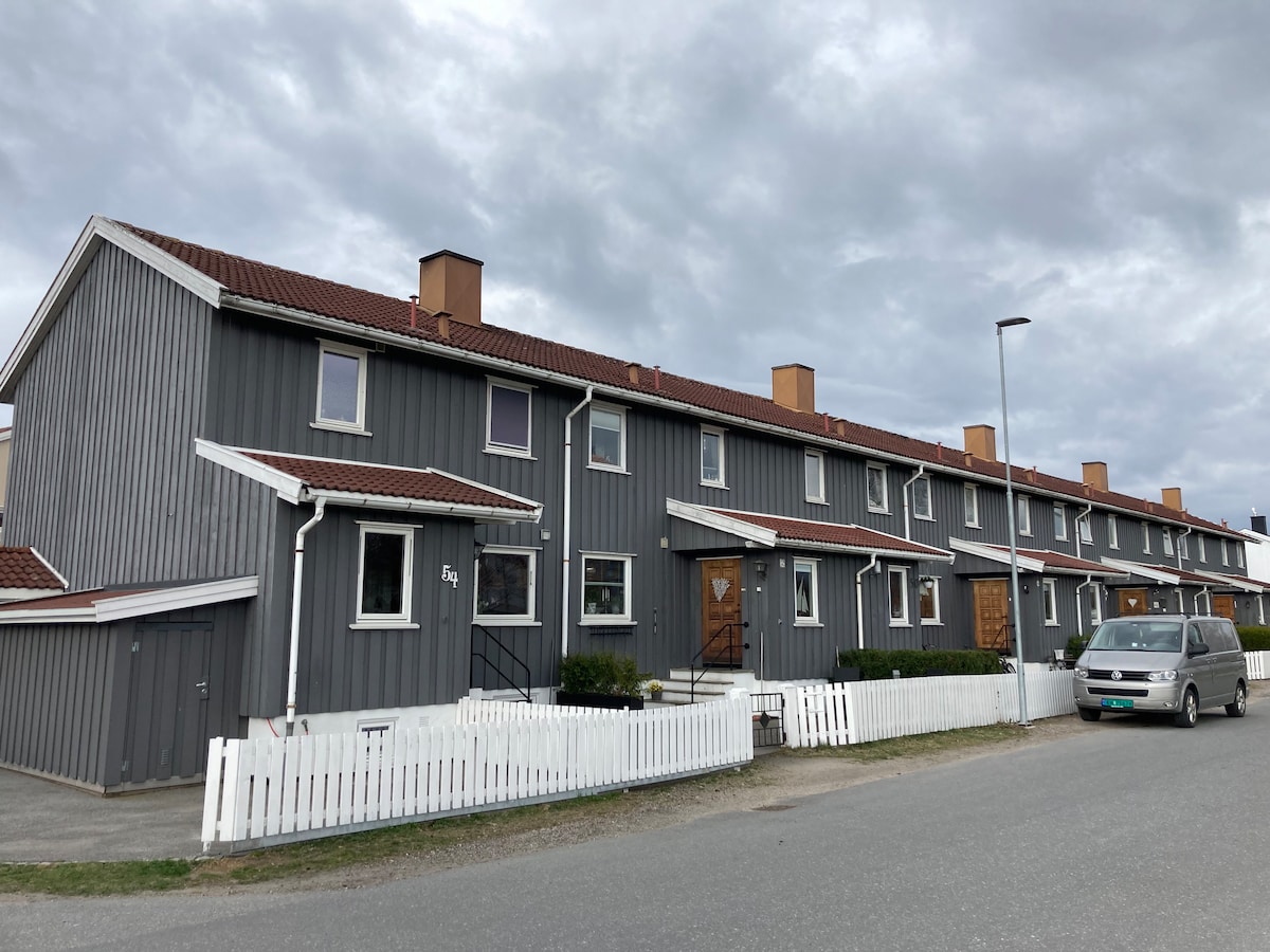 A long row of modern gray townhouses is seen from the street, each featuring multiple windows and a sloped roof. A white picket fence runs along the front, with a manicured lawn visible. Parking is available alongside the houses, with a vehicle parked nearby.