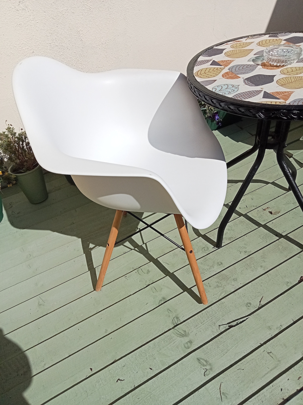 A white chair with a curved design is positioned beside a circular table featuring a patterned tabletop. The chair is supported by wooden legs on a wooden decking surface. Green foliage is visible in the background, enhancing the outdoor setting.