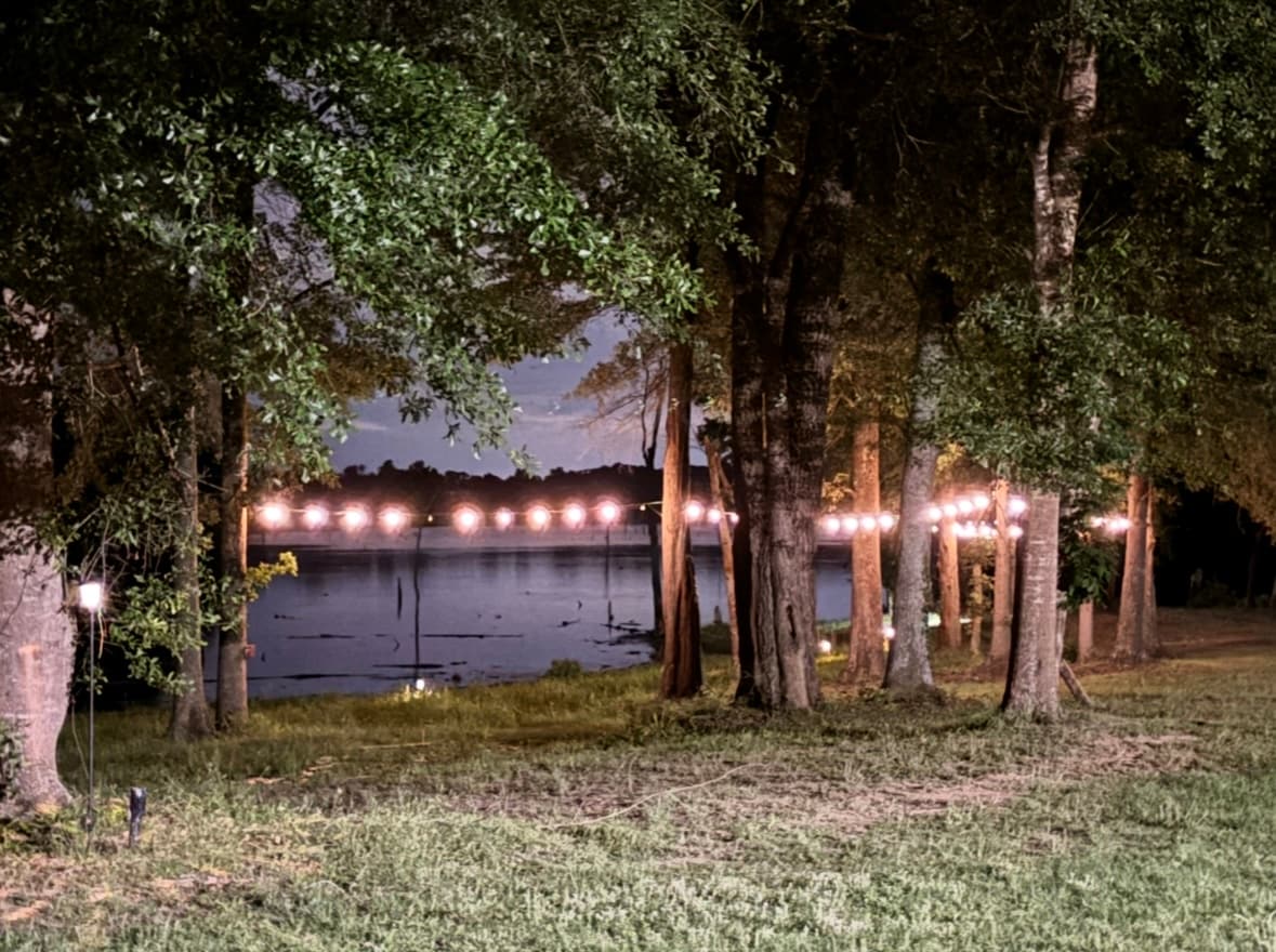 Soft lighting is strung along trees leading to the water's edge, creating a pleasant ambiance. The lake is visible in the background, framed by lush greenery, offering a serene setting for evening relaxation.