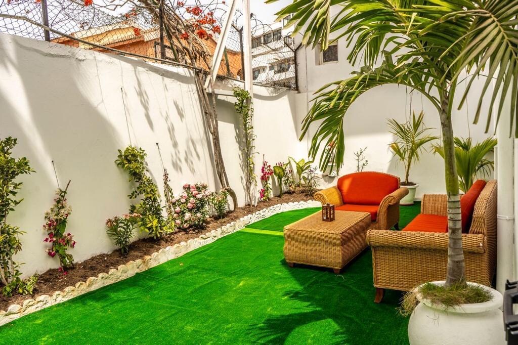 A private garden is presented with vibrant artificial grass, framed by neatly arranged plants and flowers along the walls. A comfortable seating area features a woven sofa with orange cushions, complemented by a small table. Sunlight illuminates the space, enhancing its serene atmosphere.