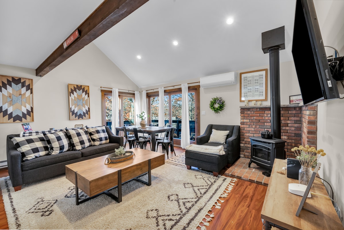 A welcoming living area is highlighted by large windows that invite natural light. A dark sofa is complemented by patterned cushions and a geometric rug. A wooden coffee table sits in the center, and a dining area with black chairs is visible in the background.