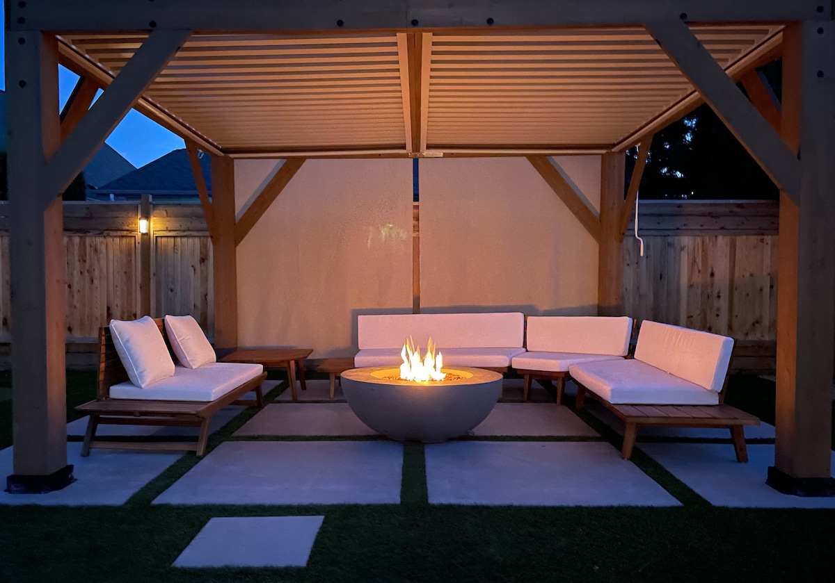 A covered outdoor seating area features modern, plush lounge seating arranged around a circular fire pit. Soft illumination highlights the space, while privacy curtains provide a cozy backdrop. The surrounding area consists of neatly maintained grass and a concrete patio, enhancing the inviting setting.