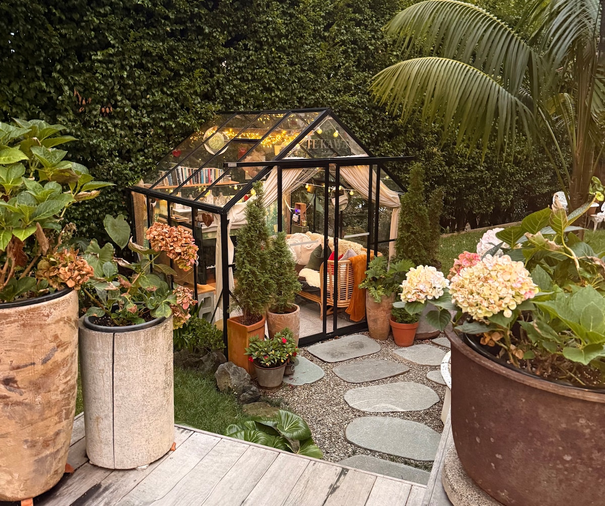A charming glasshouse nestled in a lush garden, surrounded by various potted plants and flowers. The glasshouse features inviting lighting and comfortable seating, creating a serene retreat. Exposed stone paths and greenery lead towards the entrance, enhancing the tranquil atmosphere.