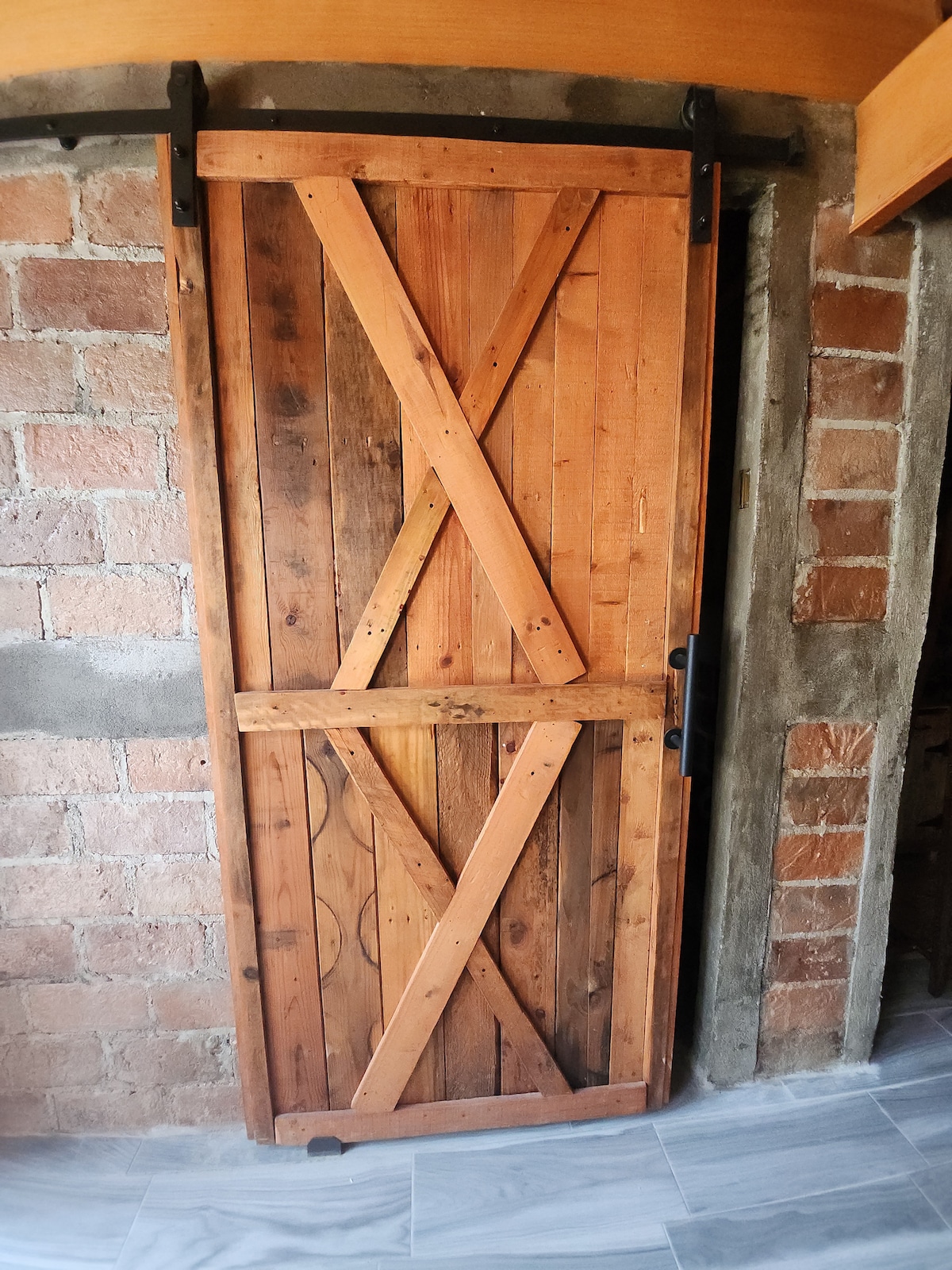 A wooden barn-style door is positioned against a brick wall, displaying a rustic charm with its cross-beam design. The natural wood grain and variations in color add texture, while a sleek black handle contrasts against the warm tones of the door.