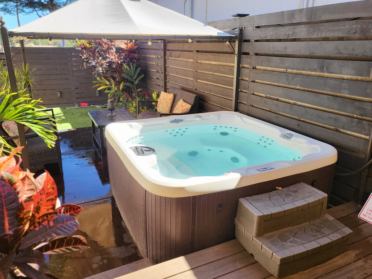 A private hot tub is nestled in an enclosed outdoor space, surrounded by tropical plants and wooden fencing. Comfortable seating arrangements are nearby, creating a serene atmosphere. Steps lead up to the hot tub, inviting relaxation under the clear sky.