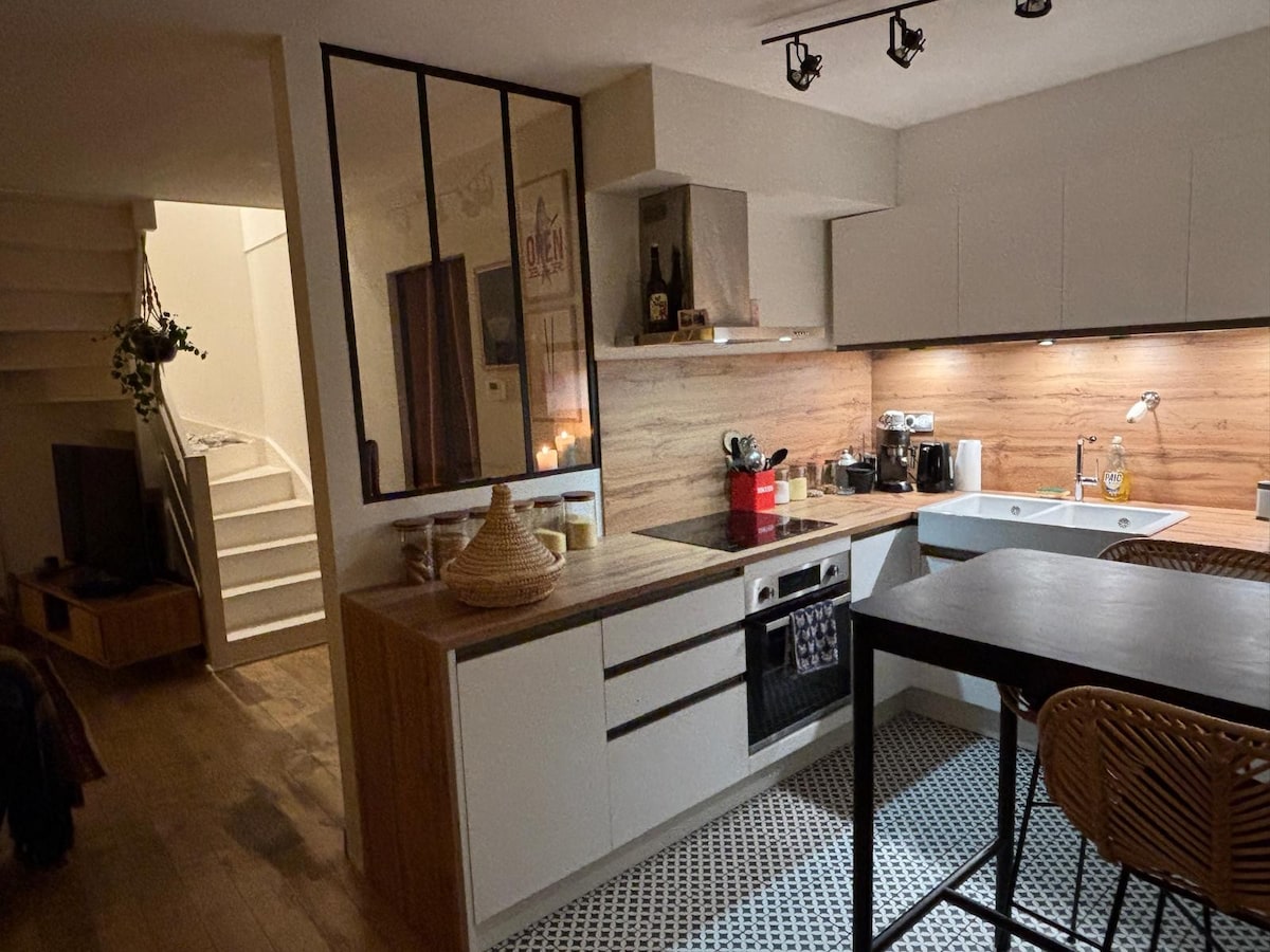 The kitchen area features modern cabinetry with wood accents and a sleek countertop. A small dining table with stools is positioned nearby, while natural light is provided through a window. The open layout connects seamlessly with the living space visible in the background.