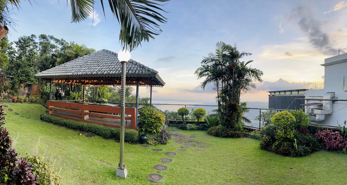 A shared gazebo is set amidst a spacious garden featuring well-maintained greenery and colorful plants. The area is bordered by a paved path leading through the garden, with a distant view of a body of water under a cloudy sky.