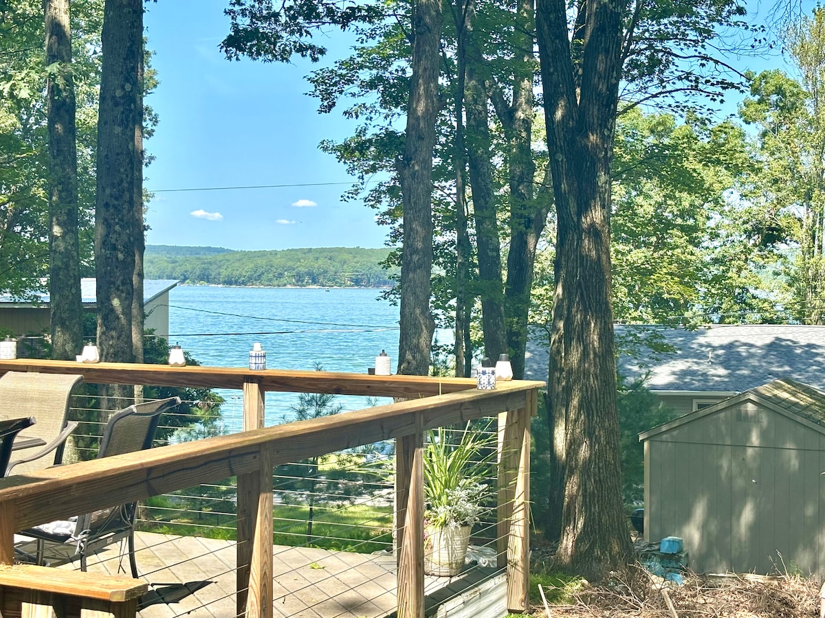 A wooden deck overlooks a serene lake, framed by tall trees and greenery. Comfortable outdoor furniture is arranged on the deck, providing a welcoming space to relax. The gentle ripples on the water reflect the clear blue sky, enhancing the tranquil atmosphere of the setting.