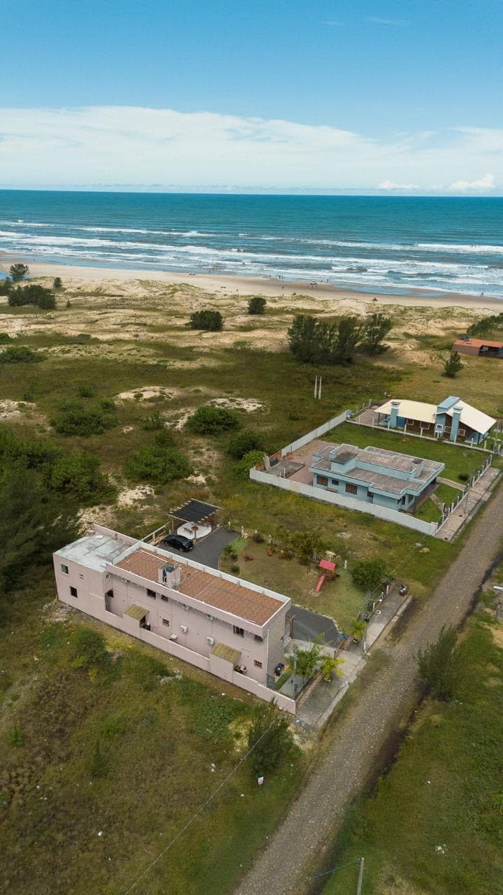 An aerial view captures a serene coastal landscape featuring pastel-colored buildings set against the beach and ocean. The expanse of sandy shore is bordered by greenery, while paths and structures indicate a quiet, inviting area close to the water.