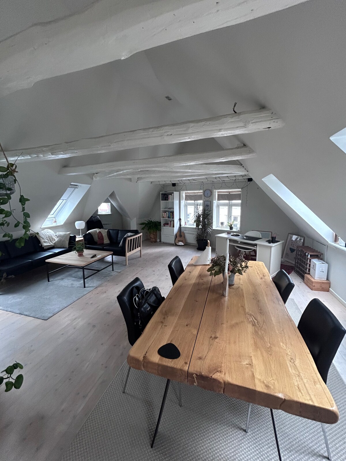 An open living area features a large wooden dining table surrounded by black chairs. A cozy seating arrangement is positioned across from the table, with a dark sofa and armchairs near the windows. Natural light floods the space through slanted ceilings and multiple skylights.