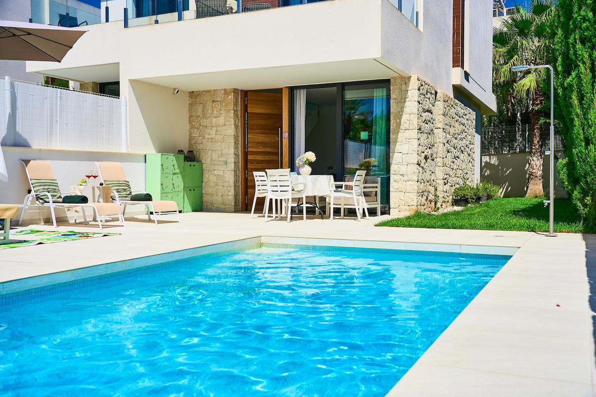 A private swimming pool is surrounded by a spacious patio area with sun loungers and a dining table. The clean lines of the modern villa are visible, and a lush garden is present in the background, providing a serene outdoor space.