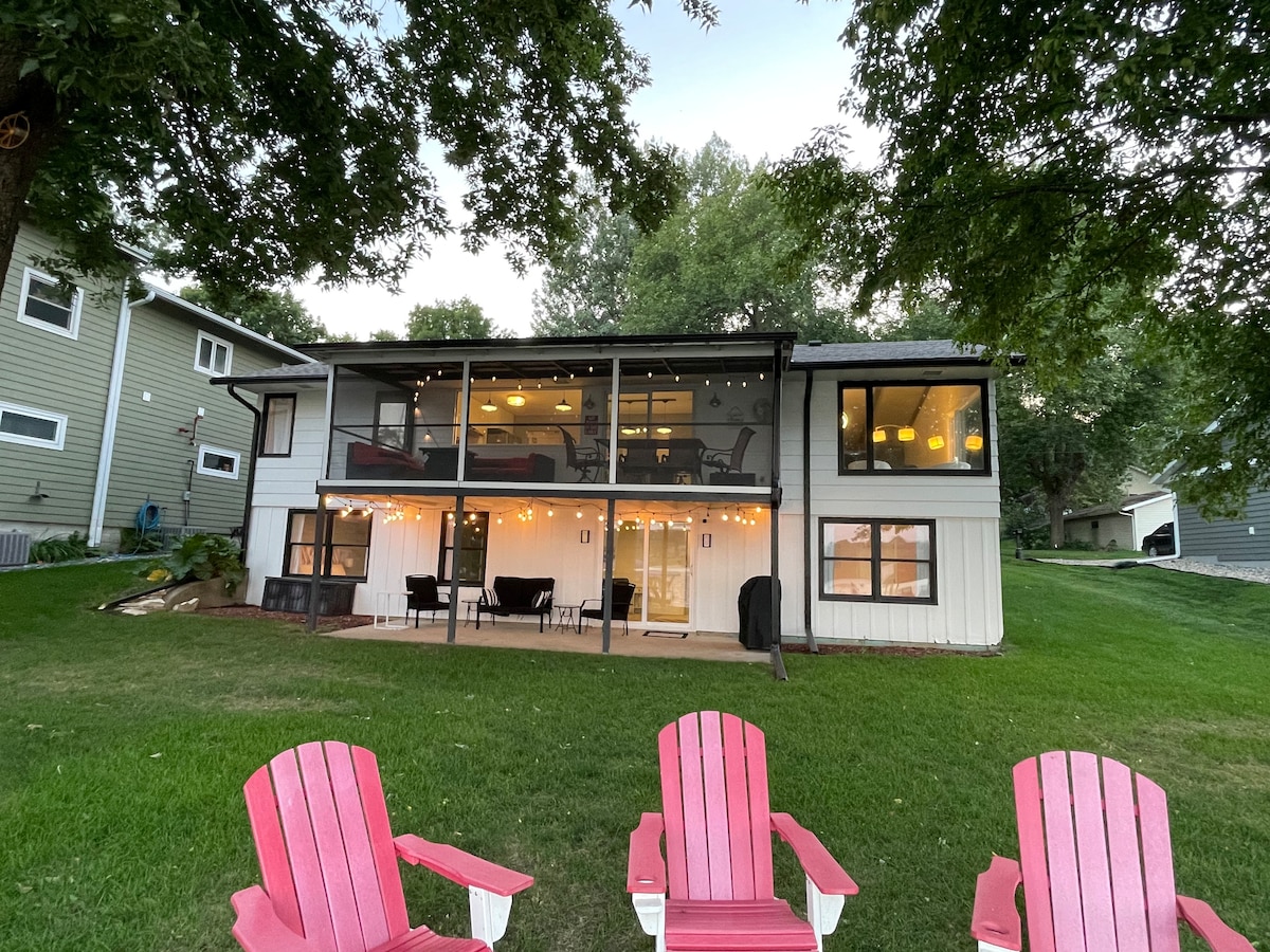 The back view of the lakefront home showcases multiple levels with a screened-in deck, illuminated by warm lights. A spacious backyard features vibrant green grass and six red Adirondack chairs arranged in front, inviting relaxation and outdoor gatherings.