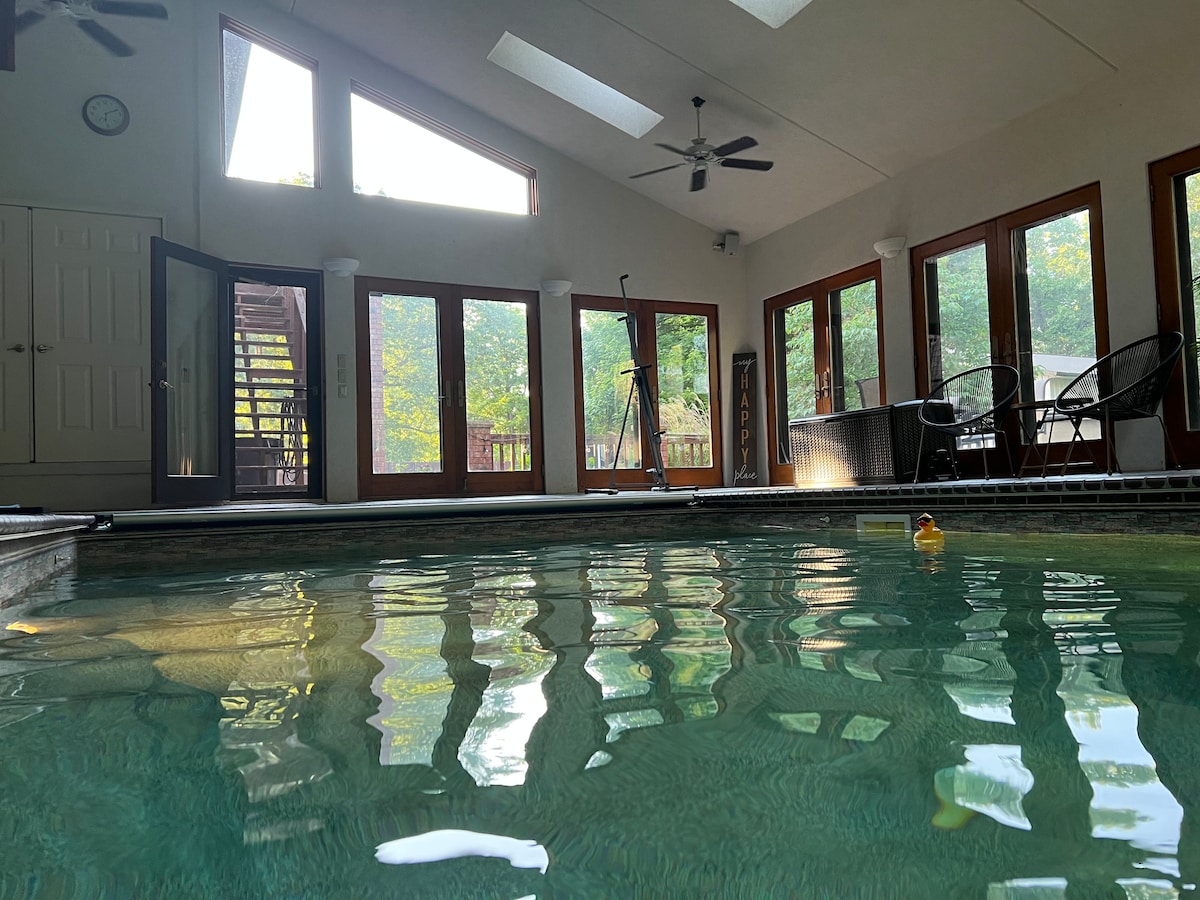 An indoor swimming pool area is bathed in natural light from large windows and skylights. The water's surface reflects the surrounding features, including a small rubber duck floating nearby. Comfortable seating is arranged by the windows, offering views of the landscaped outdoor space.