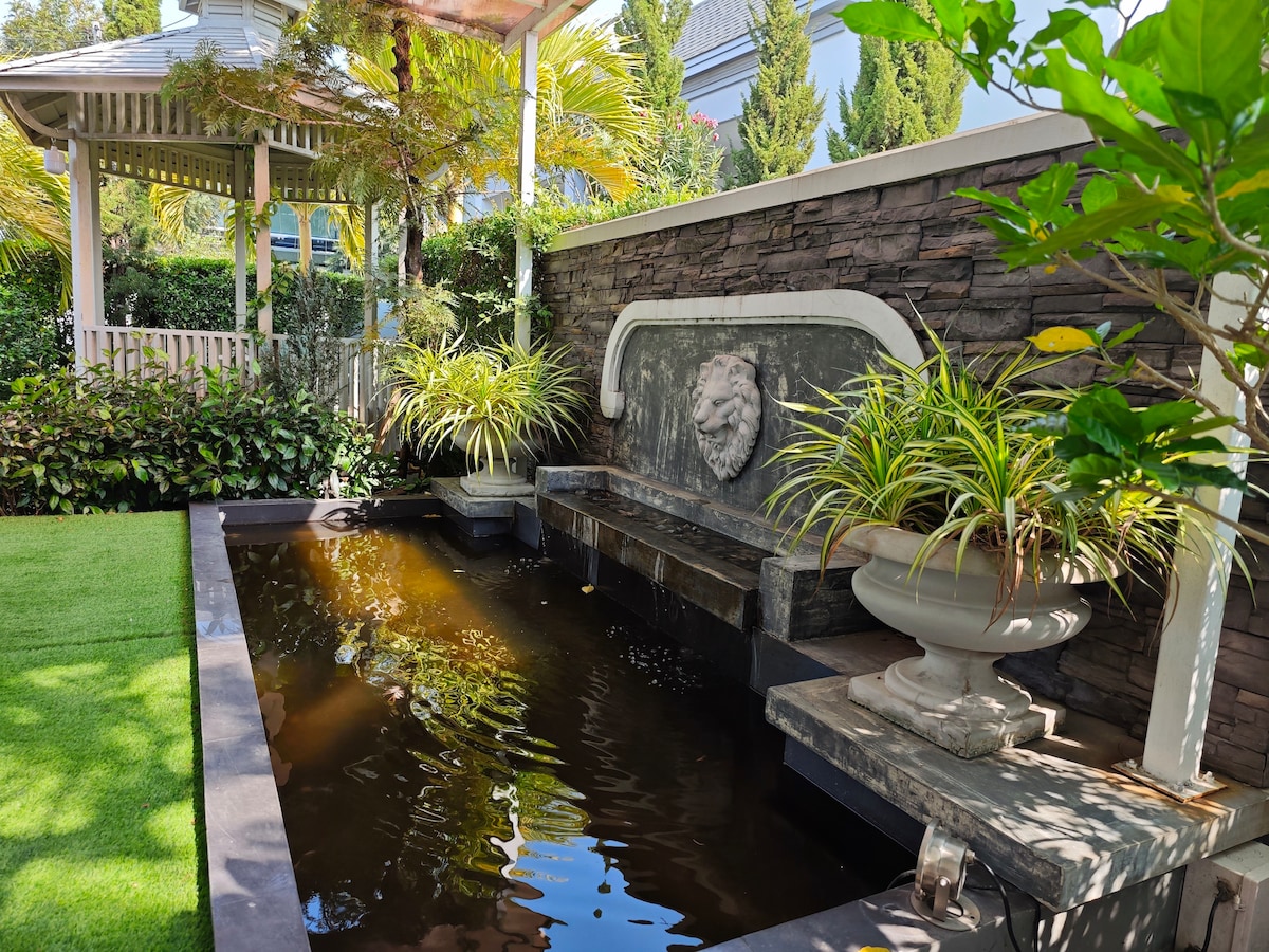 A serene water feature is set against a textured stone wall, complete with a carved lion's head. Lush greenery and ornamental plants frame the area, while a stylized seating space is positioned nearby, enhancing the calm ambiance.