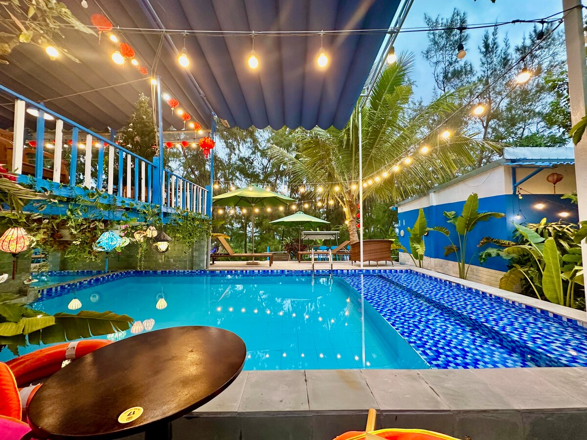 A vibrant swimming pool is framed by colorful lanterns and lush greenery. Sun loungers and umbrellas provide relaxation spots around the pool, while string lights create a warm ambiance in the evening. The area is designed for leisure and enjoyment in a natural setting.