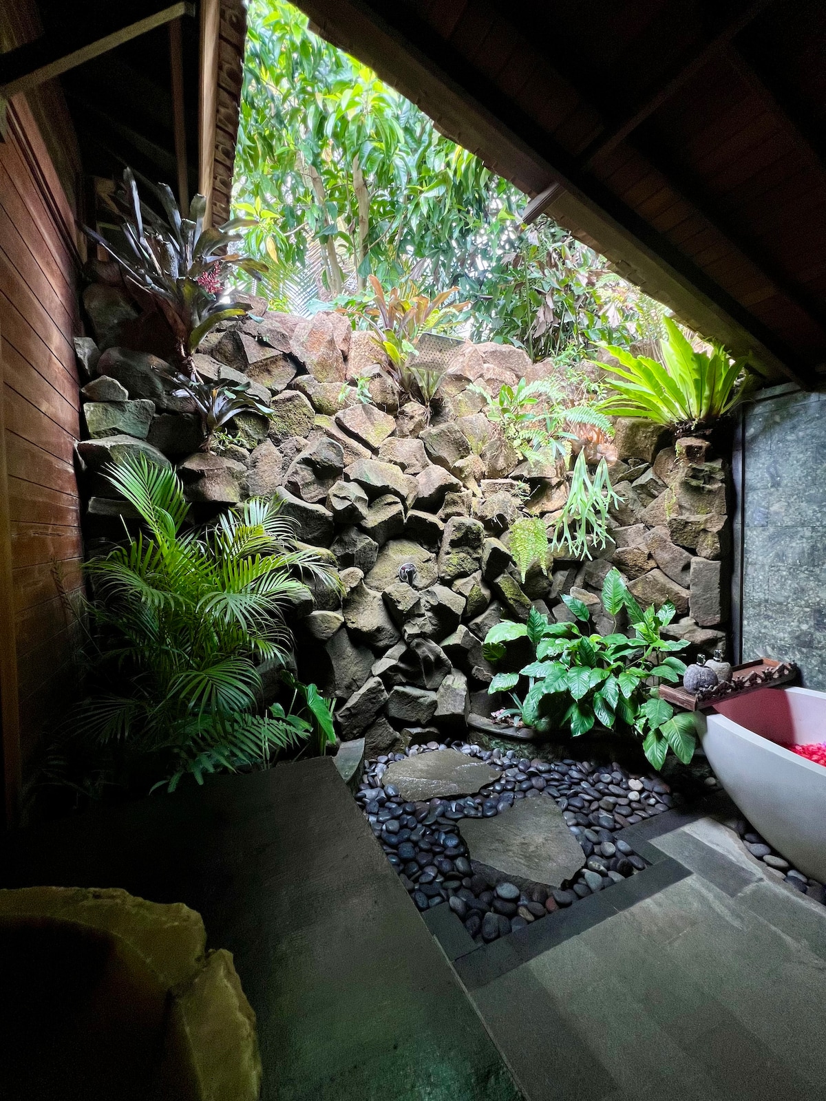A semi-open bathroom features natural stone walls, enhancing the connection to nature. Lush green plants and a small pebble pathway contribute to a serene atmosphere. A freestanding bathtub is positioned nearby, allowing for a tranquil bathing experience.