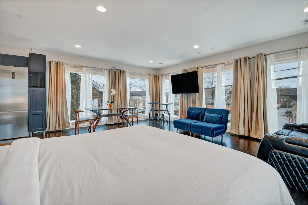 Bright and inviting interior showcased through panoramic windows. Elegant furnishings include a spacious bed with crisp linens, a modern sofa, and a wooden dining table. A flat-screen TV is mounted on the wall, while soft fabric drapes complement the serene atmosphere.